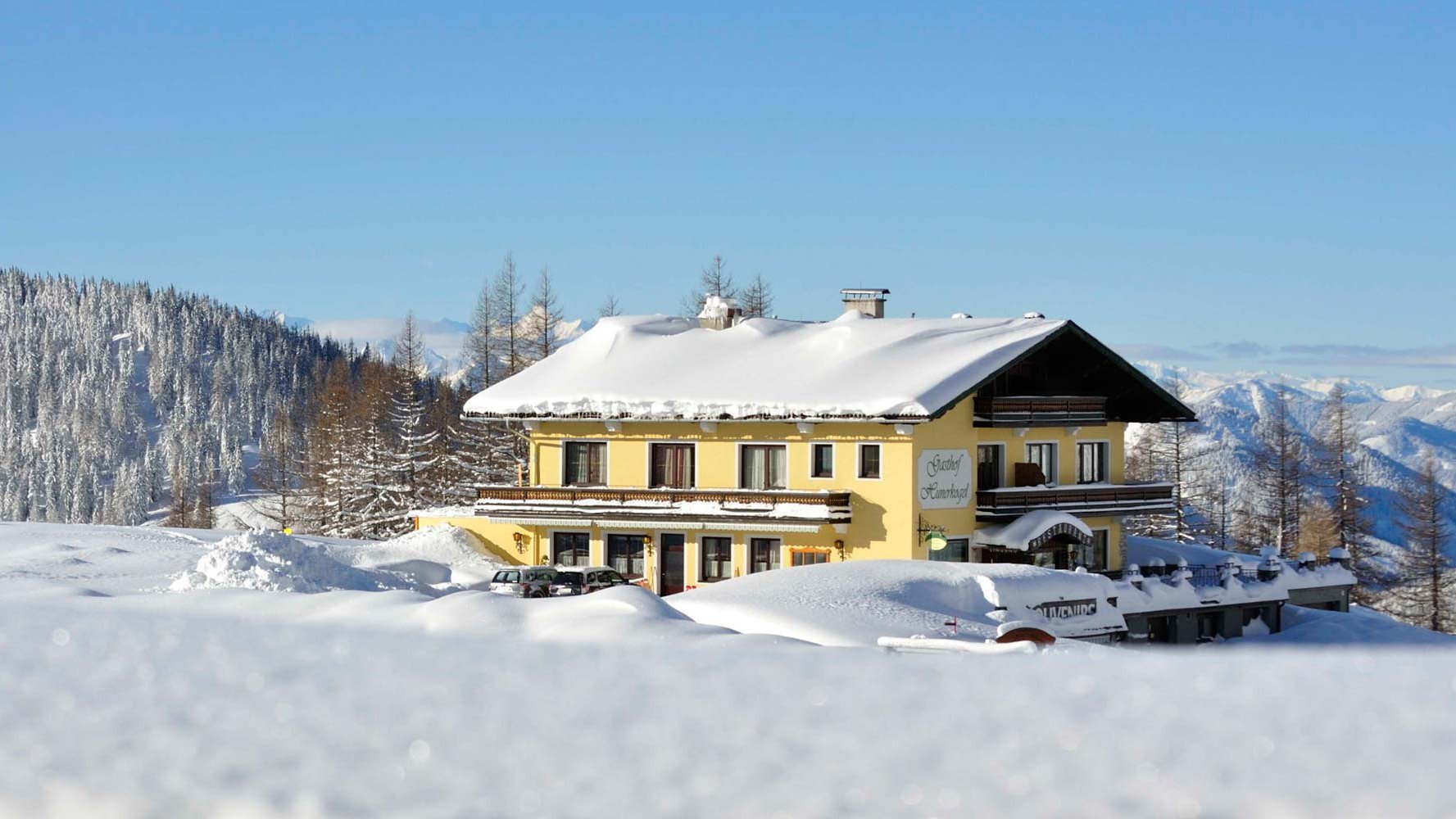Alpenidylle am Gasthof Hunerkogel Berglodge in verschneiter Landschaft mit Kiefern und Bergen im Hintergrund unter blauem Himmel